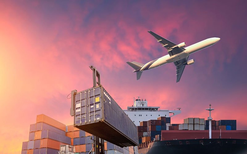 A cargo airplane flying over a container ship and stacked shipping containers at a port during a vibrant sunset, representing multimodal shipping from China to the UAE.