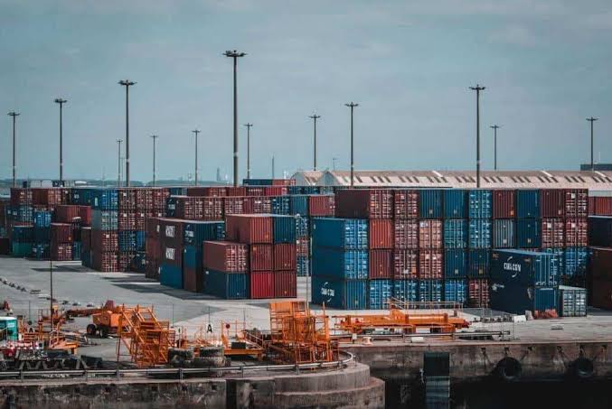 A large container yard filled with stacked blue, red, and brown shipping containers, with orange port machinery in the foreground and tall lighting poles under a cloudy sky.